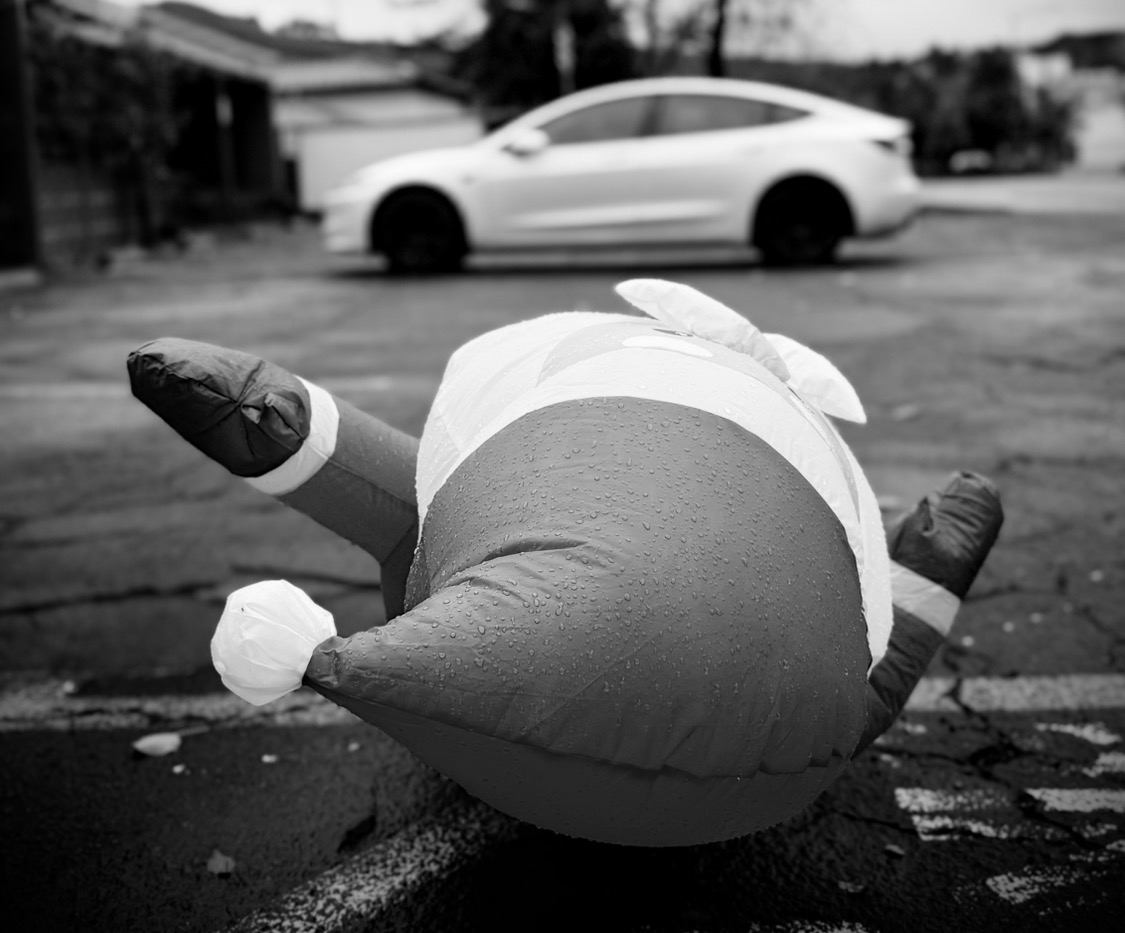 A deflated inflatable Santa Claus lies face-down on rain-soaked pavement in a Moraga, California parking lot, arms splayed, with a white car in the background